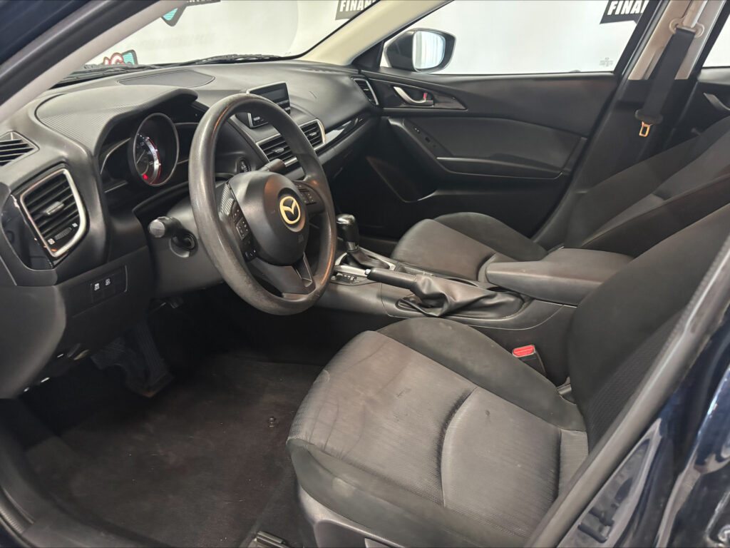 Interior of a dark car showing the steering wheel with a logo, dashboard, center display, and front seats with a seatbelt visible.”, “Left-side view of a car’s cabin featuring the steering wheel, multimedia screen, air vents, and dark fabric seats.”, “Front passenger area with door panel, seatbelt, and center console in a dark, modern car interior.”