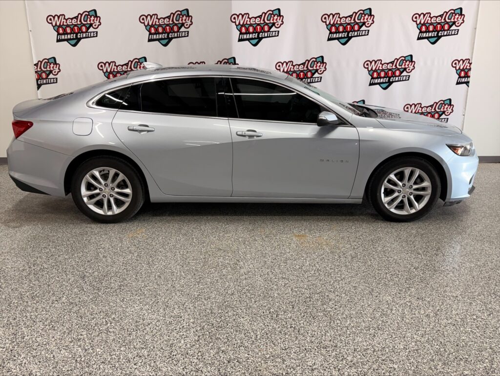 Silver Chevrolet Malibu sedan shown in profile inside a showroom with Wheel City Auto banners in the background.
