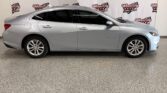 Silver Chevrolet Malibu sedan shown in profile inside a showroom with Wheel City Auto banners in the background.