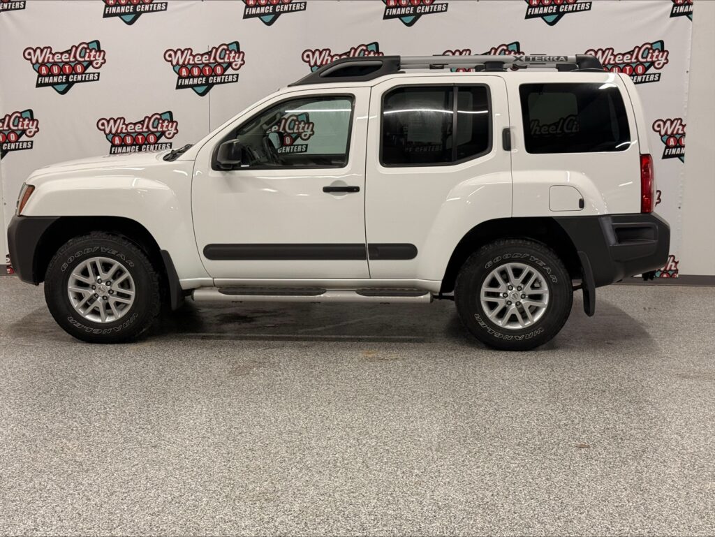 White Nissan Xterra SUV with roof rack and running boards, indoors in front of a Wheel City Auto backdrop.