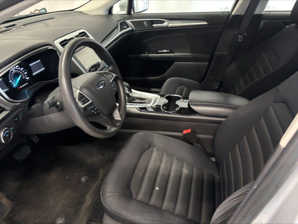 Interior of a car cockpit: steering wheel, center console with cup holders, and black front seats viewed from the passenger side.