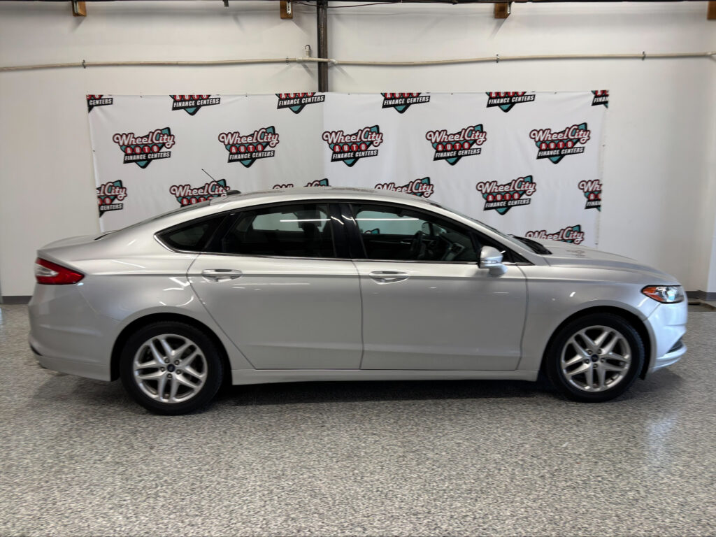 Side profile of a silver sedan parked indoors in front of a white backdrop with 'Wheel City Auto Finance Centers' logos repeated across the banner.