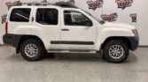 White Nissan Xterra/SUV shown in a dealership showroom, side profile with roof rack and running boards.