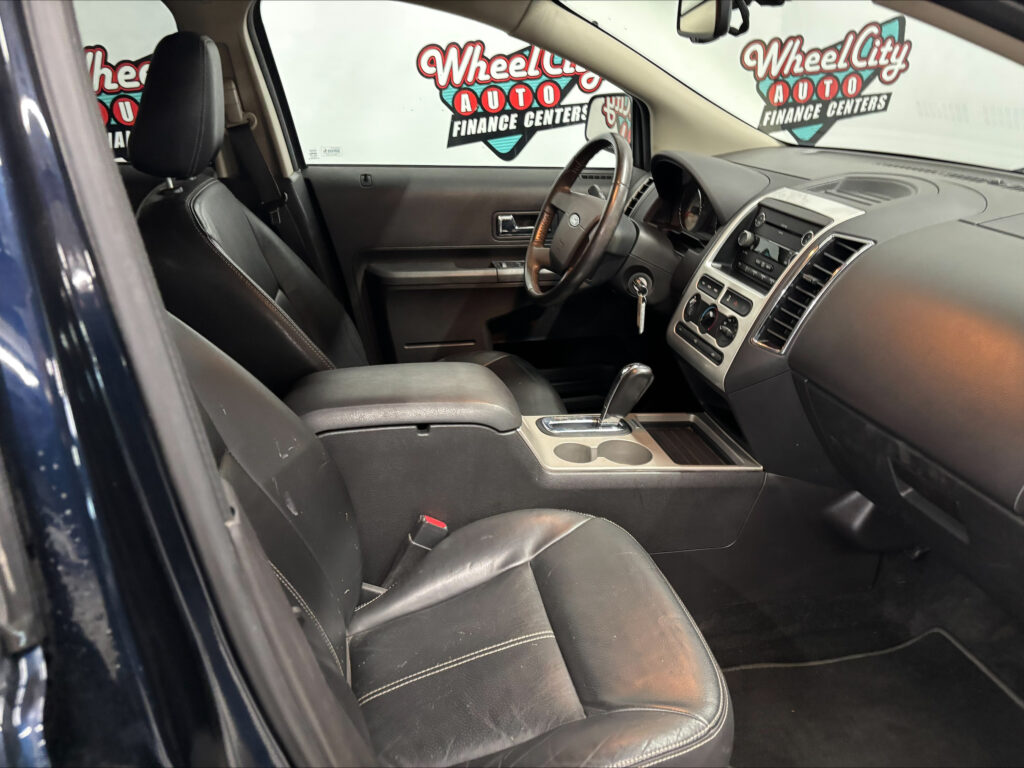 Interior of a black leather SUV showing steering wheel, gear shift, and center console; dealership banner visible through the windows with Wheel City Auto Finance Centers logos.