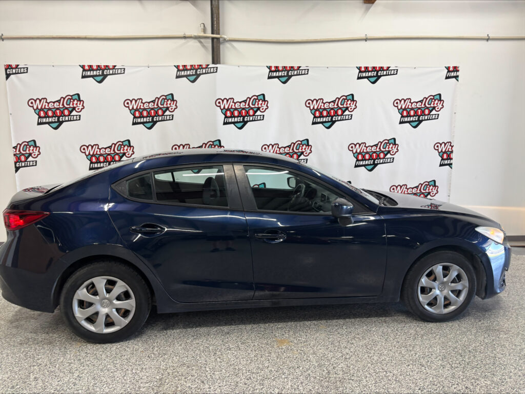 Blue sedan parked indoors in front of a White backdrop with Wheel City Auto Finance Centers logos repeated across it.