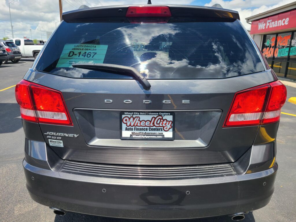 Rear view of a gray Dodge Journey SUV at a car dealership, with a green D-1467 tag visible in the rear window and red taillights showing.