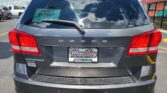 Rear view of a gray Dodge Journey SUV at a car dealership, with a green D-1467 tag visible in the rear window and red taillights showing.