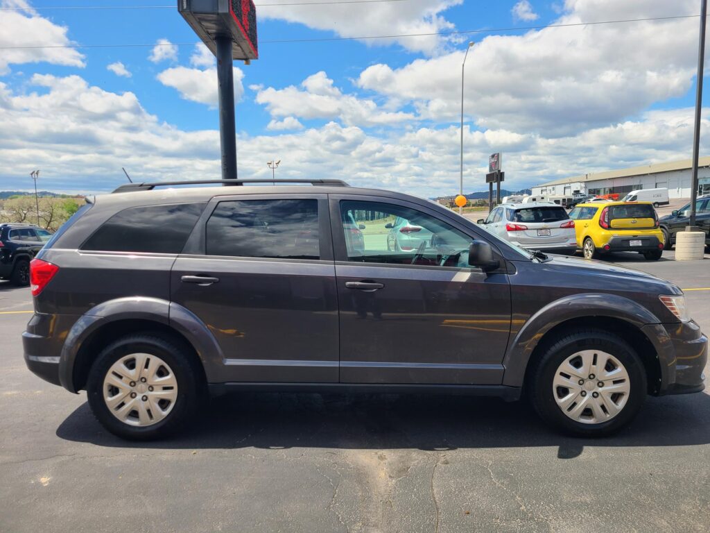 Side view of a dark gray SUV parked in a lot, with blue sky and fluffy white clouds in the background and other vehicles nearby.