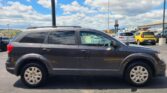 Side view of a dark gray SUV parked in a lot, with blue sky and fluffy white clouds in the background and other vehicles nearby.