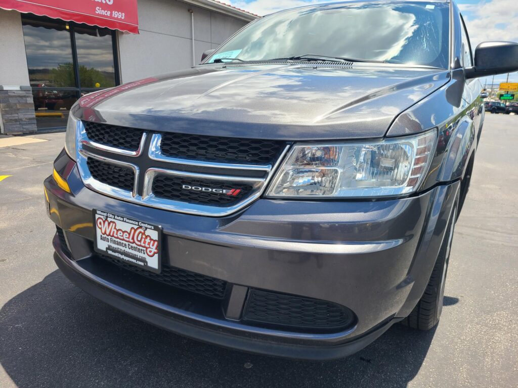 Close-up of a gray Dodge front end at a car dealership, hood reflecting a cloudy sky; Wheel City license plate frame visible in foreground.
