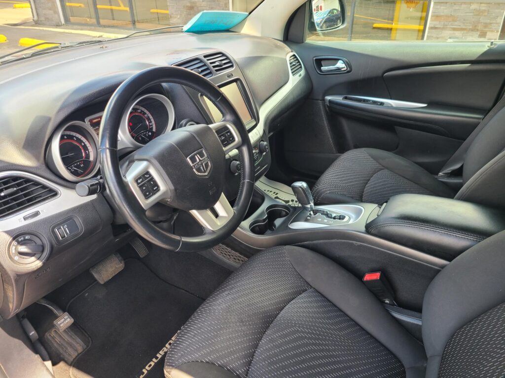 Driver's-seat view of a Dodge SUV interior: steering wheel, instrument cluster, center touchscreen, and gear shifter visible amid black fabric seats.