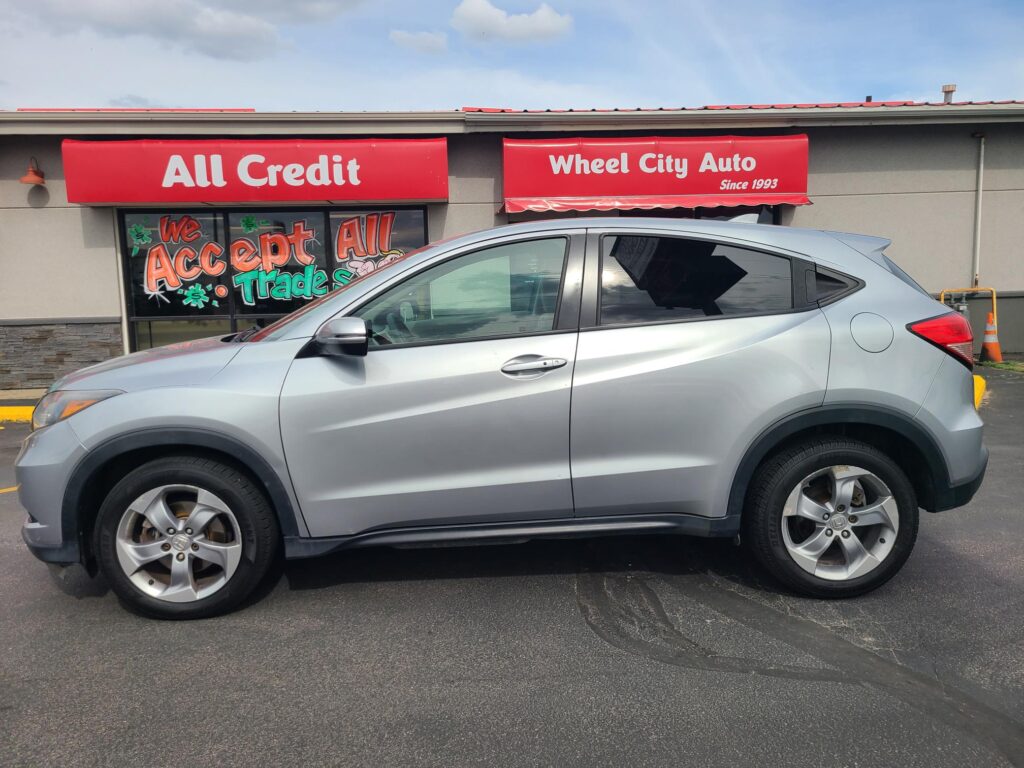 Silver SUV parked in front of a storefront with red All Credit and Wheel City Auto signs; car partially blocks the window display saying We Accept All Trade.