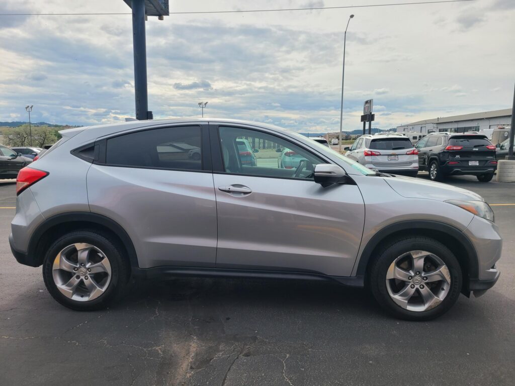 Side view of a silver Honda SUV parked in a busy lot, with other vehicles and a dealership building in the background under cloudy skies.