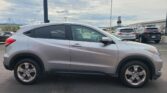 Side view of a silver Honda SUV parked in a busy lot, with other vehicles and a dealership building in the background under cloudy skies.