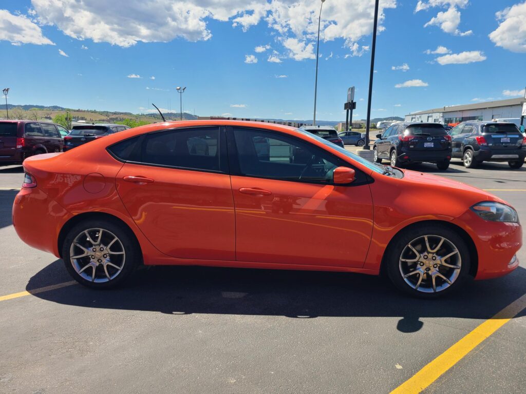 Bright orange sedan parked in a busy parking lot on a sunny day, with other cars and a building in the background.