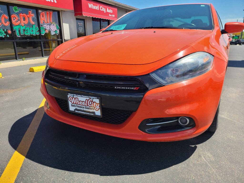 Orange Dodge SUV parked in front of Wheel City Auto dealership, storefront windows and red signage visible in the background.