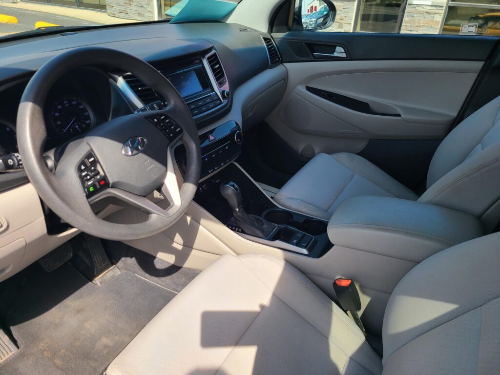 Interior of a car cabin focusing on the driver's seat, steering wheel, center console, and front passenger seat in light gray upholstery