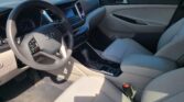 Interior of a car cabin focusing on the driver's seat, steering wheel, center console, and front passenger seat in light gray upholstery