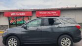 Gray Chevrolet Equinox SUV parked in front of a used-car storefront with red 'All Credit' and 'Wheel City Auto' signs above the entrance.