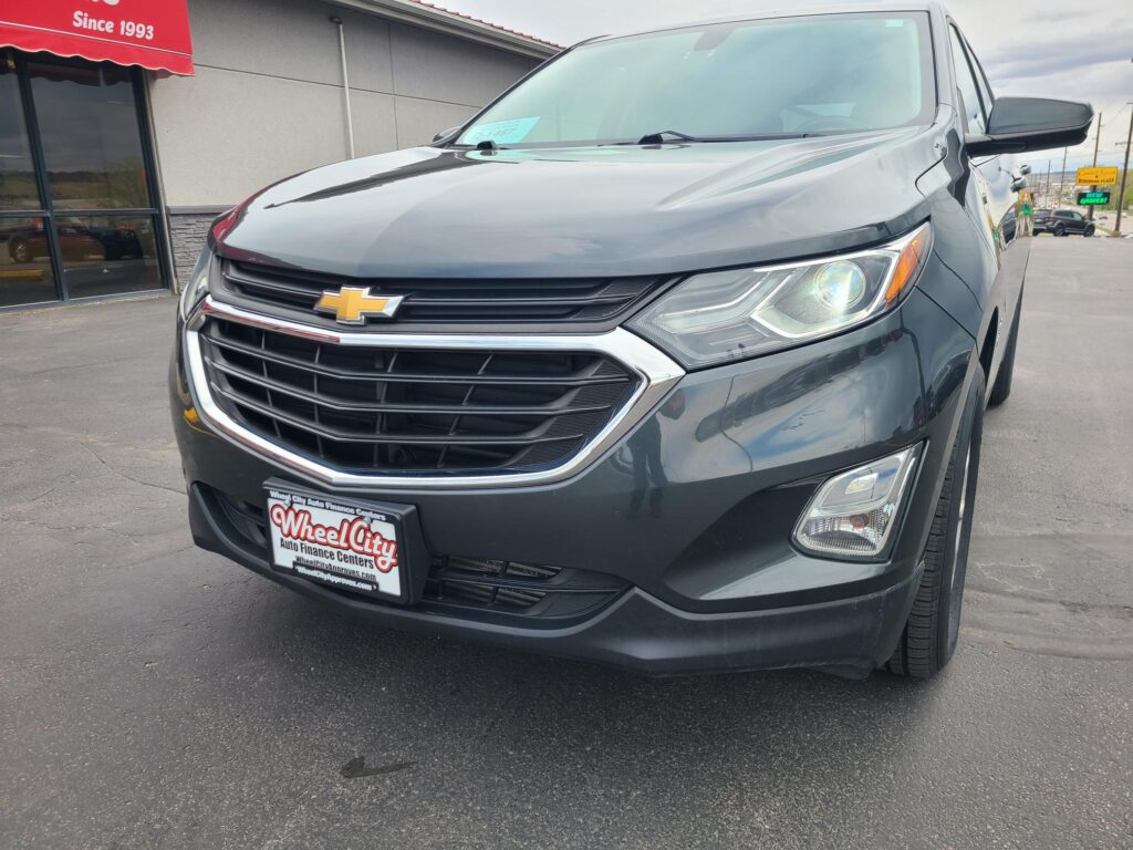 Close-up of a gray Chevrolet SUV parked outside a dealership, showing the front grille, headlight, and Chevy emblem with a Wheel City Auto Finance Centers license plate frame visible.
