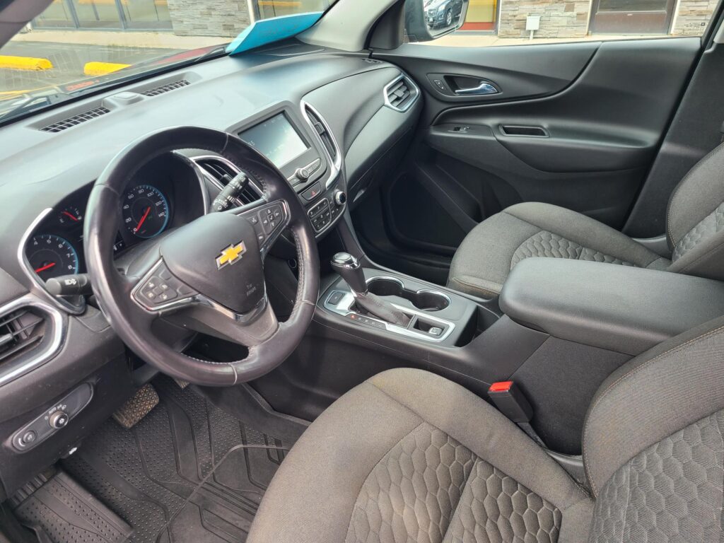 View of the car's black interior: steering wheel, center touchscreen, gear selector, and front passenger seat in front of you