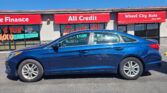 Blue sedan parked in front of a storefront auto dealership with red signs reading 'All Credit' and 'Wheel City Auto' under a clear blue sky