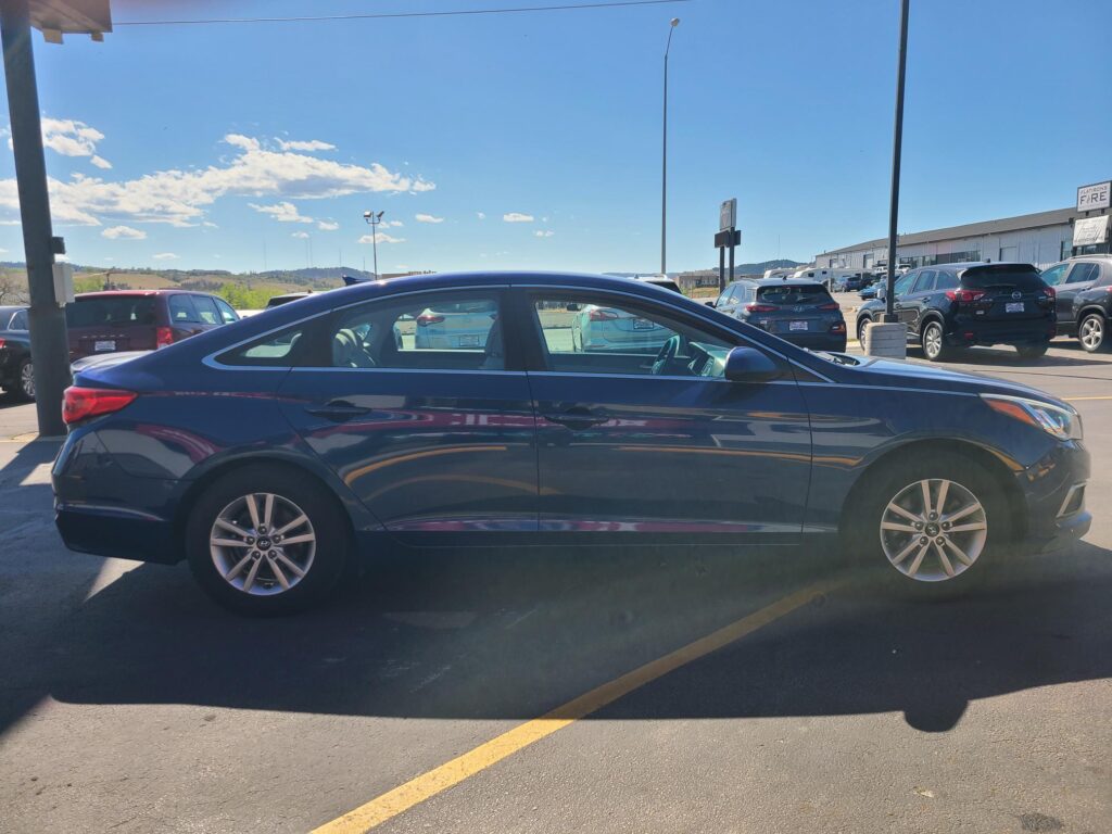 Blue car in foreground parked in a lot, with other vehicles and a low, clear blue sky in the background.