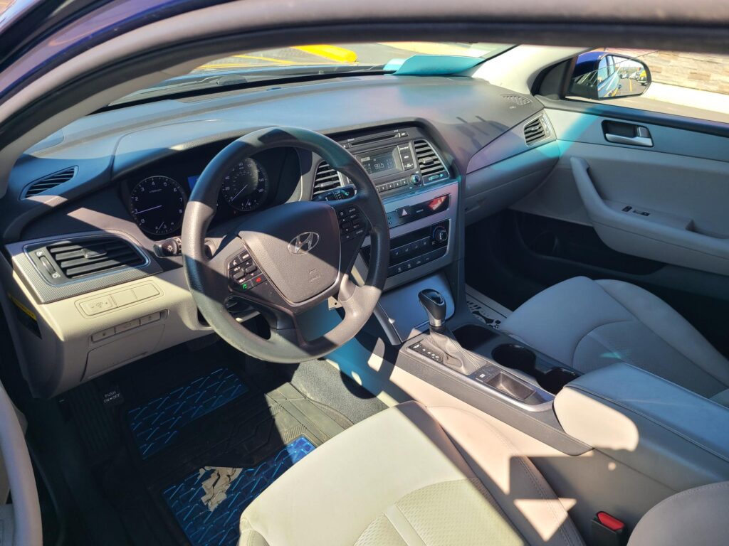 Front interior of a car showing the steering wheel, dashboard, center console, and light gray seats in daylight.
