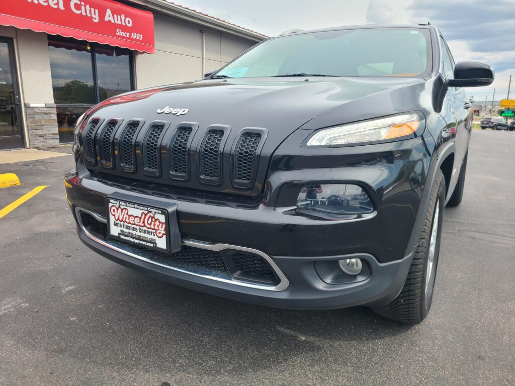 Close-up view of a dark gray Jeep SUV parked in front of a Wheel City Auto dealership, featuring the seven-slot grille and modern headlights.