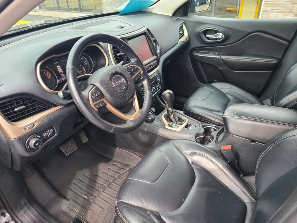 Front interior of a black leather Jeep, featuring the steering wheel, center touchscreen, gear shifter, and leather seats.
