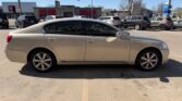 Beige four‑door sedan parked in a commercial parking lot, side view with tinted windows and alloy wheels; other cars and storefronts visible in the background.