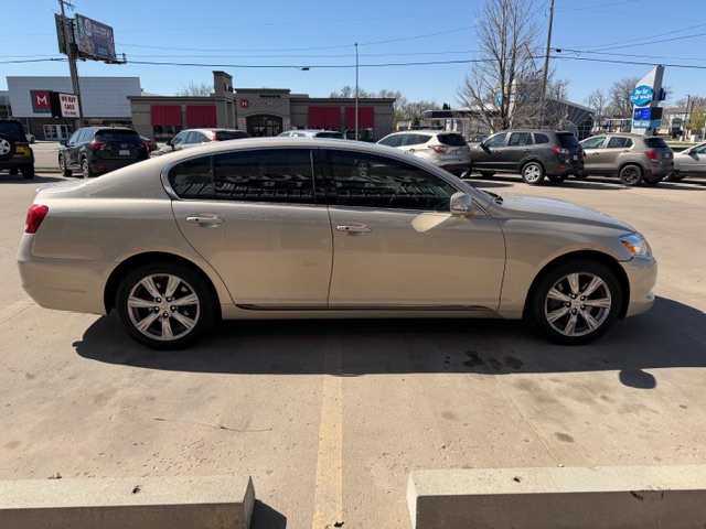 Beige four‑door sedan parked in a commercial parking lot, side view with tinted windows and alloy wheels; other cars and storefronts visible in the background.