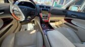 Interior view of a beige leather Lexus sedan: steering wheel on the left, wood-trim center console with touchscreen, gear shifter, and front seats bathed in sunlight from the windows.