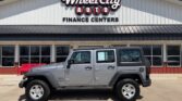Silver Jeep Wrangler parked in front of Wheel City Auto Finance Centers storefront on a sunny day.