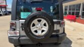 Rear view of a gray Jeep Wrangler with a spare tire mounted on the back, a Firestone Destination AT tire on the spare, window sticker visible through the rear glass, and a dealership building with a red bench on the right.