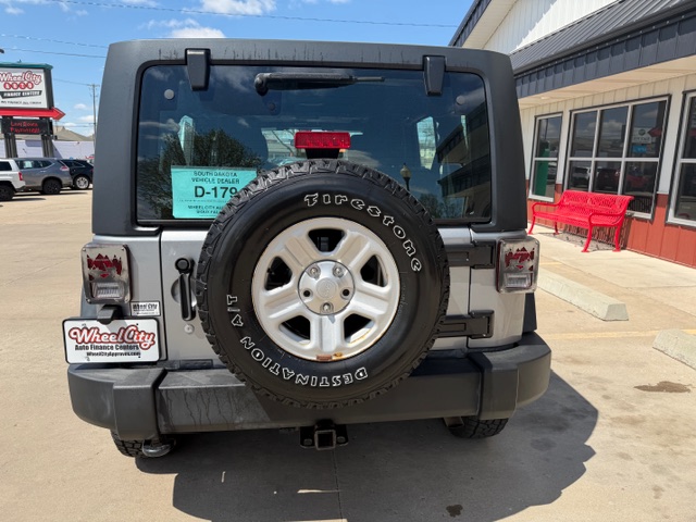 Rear view of a gray Jeep Wrangler with a spare tire mounted on the back, a Firestone Destination AT tire on the spare, window sticker visible through the rear glass, and a dealership building with a red bench on the right.