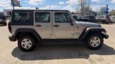 Silver Jeep Wrangler Unlimited four-door SUV with a black hard top, parked in a sunny car lot; side view showing doors, windows, and alloy wheels.