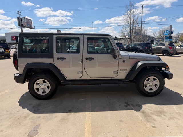 Silver Jeep Wrangler Unlimited four-door SUV with a black hard top, parked in a sunny car lot; side view showing doors, windows, and alloy wheels.