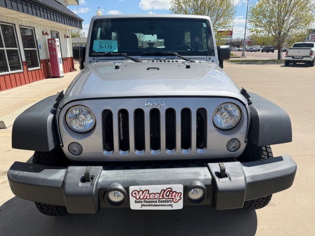 Front view of a silver Jeep Wrangler with round headlights and seven-slot grille in a dealership lot.