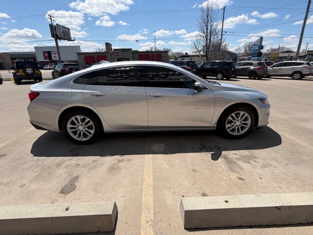 Side view of a silver mid-size sedan parked in a parking lot, with other vehicles and a strip mall in the background.