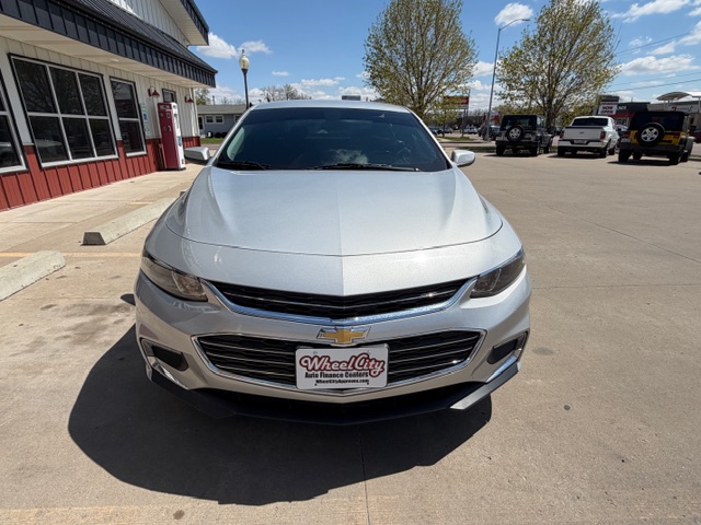 Front view of a silver Chevrolet sedan parked in a dealership lot, with a red building to the left and other vehicles in the background.