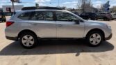 Side view of a silver Subaru Outback SUV parked in a lot, with roof rails and a row of other cars and storefronts in the background.
