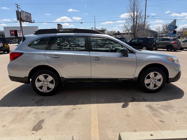 Side view of a silver Subaru Outback SUV parked in a lot, with roof rails and a row of other cars and storefronts in the background.