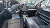 Interior of a Subaru car: steering wheel with controls, center console and infotainment screen, gear shifter, and black fabric seats; dealership lot visible through windows.