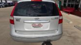 Rear view of a silver Hyundai Touring minivan at a dealership, with a Wheel City license plate frame and a red bench nearby.
