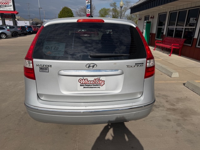 Rear view of a silver Hyundai Touring minivan at a dealership, with a Wheel City license plate frame and a red bench nearby.