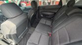 Back seat area of a dark fabric car interior, three passenger seats with headrests and seat belts visible from the rear door. Outside storefronts are seen through the windows.