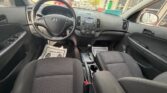 Interior of a Hyundai SUV showing black fabric seats, steering wheel, center console with radio and climate controls, gear shifter, and cup holders; dealership lot visible through the windows.