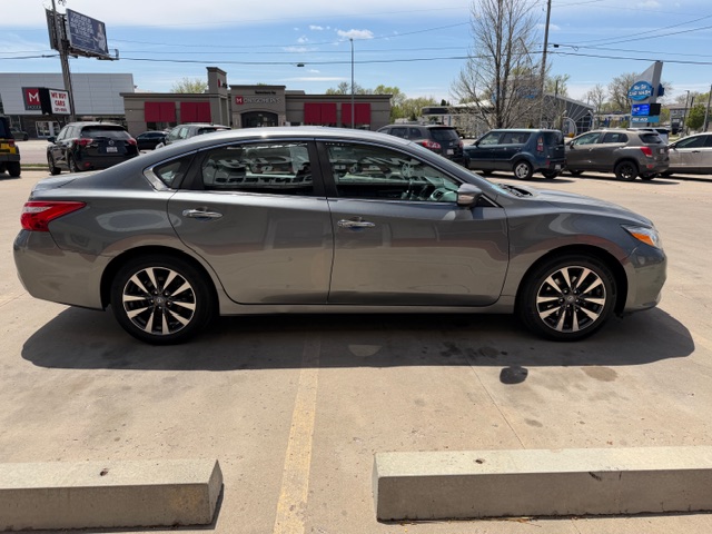 Side view of a gray four-door sedan parked in a concrete parking lot, with dealership buildings and other cars in the background.