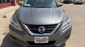 Front view of a gray Nissan sedan at a dealership, Wheel City Auto Finance Centers license frame, parking lot and buildings in the background.